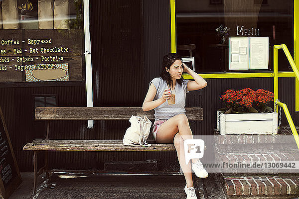 Nachdenkliche Frau trinkt Kaffee  während sie auf einer Bank im Bürgersteig-Café in der Stadt sitzt