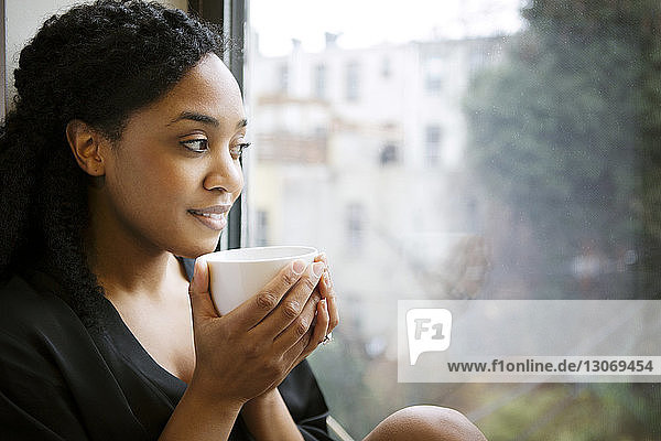 Lächelnde Frau mit Tasse  die wegschaut  während sie am Fenster sitzt