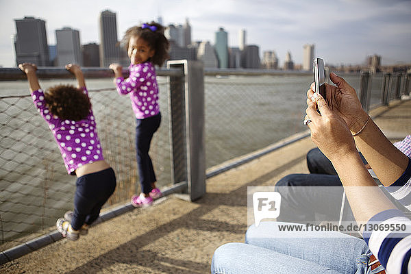 Mother using smart phone while daughters playing at railing against sea in city