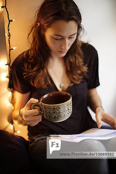 Serious woman holding coffee cup while reading book at home