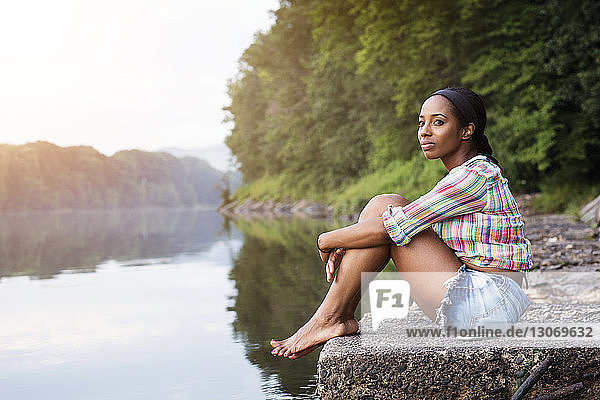 Seitenansicht einer Frau  die weg schaut  während sie auf einem Felsen gegen einen See im Wald sitzt