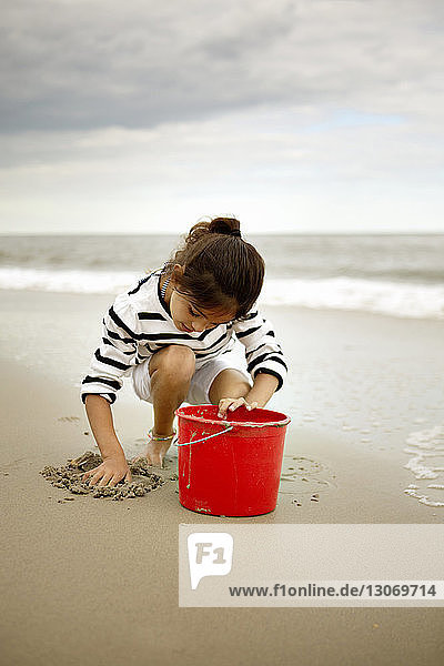 Mädchen spielt mit Sand  während sie am Strand vor bewölktem Himmel kauert