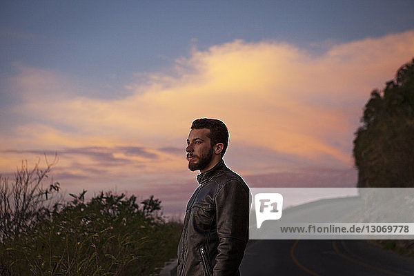Nachdenklicher Mann schaut weg  während er bei Sonnenuntergang auf der Küstenstraße steht