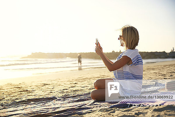 Side view of woman photographing while sitting at beach