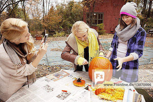Frau fotografiert  während Freunde bei Tisch einen Halloween-Kürbis schnitzen