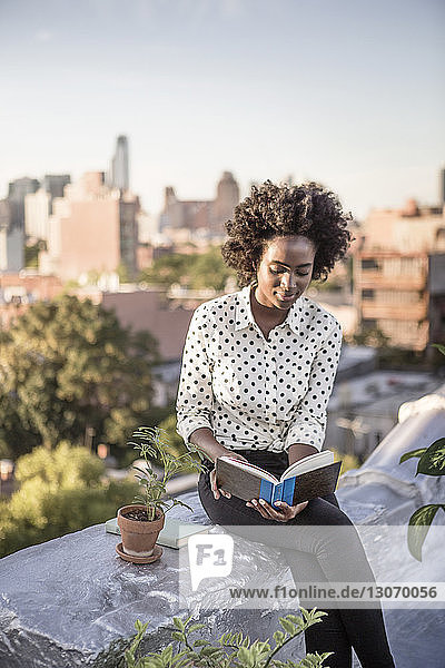 Lächelnde Frau liest Buch  während sie auf einer Gebäudeterrasse gegen den Himmel sitzt