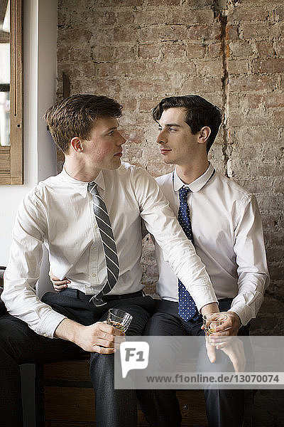Gay couple looking at each other while sitting against brick wall at home