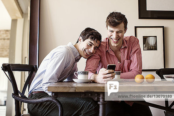 Lächelnde Schwule  die am Tisch sitzend auf ein Smartphone schauen