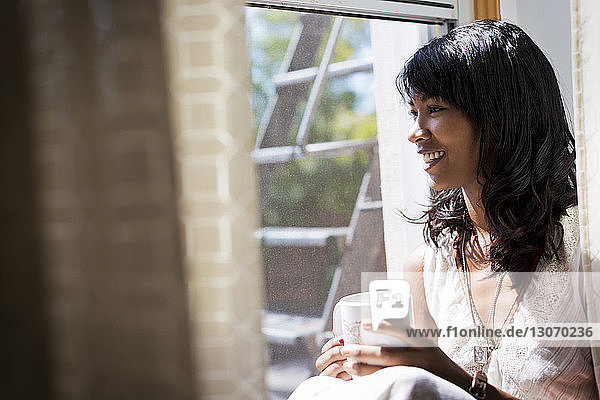 Woman with coffee cup looking away while sitting on window sill