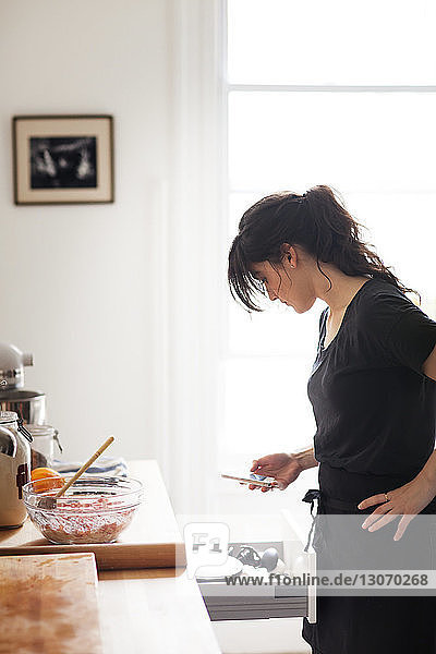 Side view of woman using smart phone while working in kitchen at home