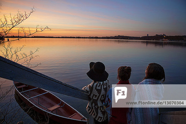 Rückansicht von Freunden  die während des Sonnenuntergangs im Winter mit einem verankerten Boot auf den See schauen