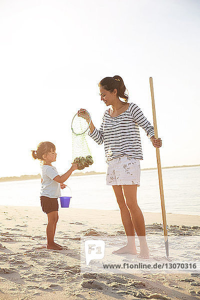 Mother and daughter with rake and clams on seashore against clear sky