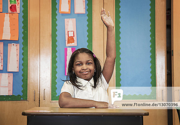 Porträt eines Mädchens  das die Hand hebt  während es am Schreibtisch im Klassenzimmer sitzt