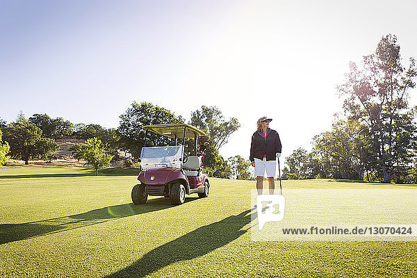 Woman looking away while standing by golf cart on field against clear sky