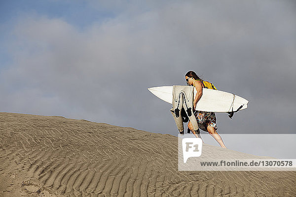 Frau in voller Länge mit Surfbrett beim Gehen auf Sand gegen den Himmel