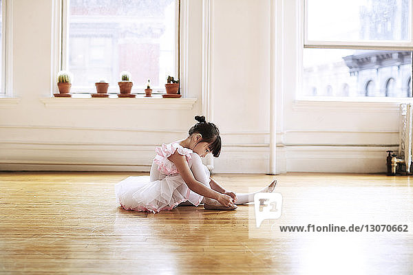Side view of girl wearing ballet shoes while sitting on hardwood floor in studio