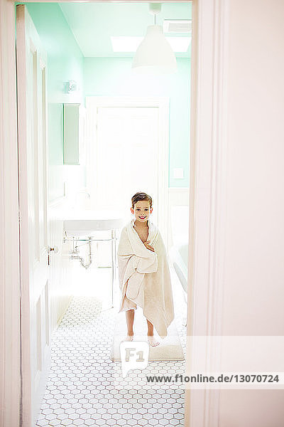 Porträt eines in ein Handtuch gewickelten Jungen im Badezimmer stehend