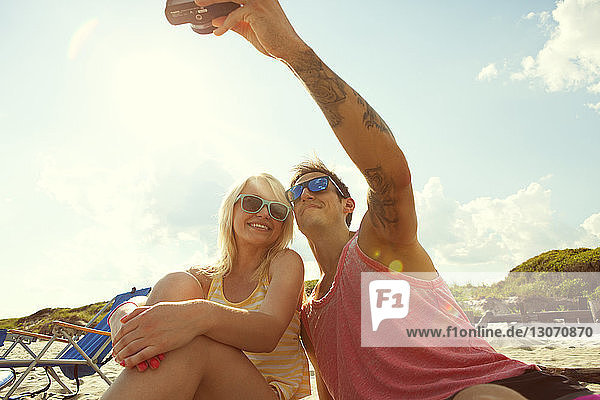 Couple taking selfie while sitting at beach against sky
