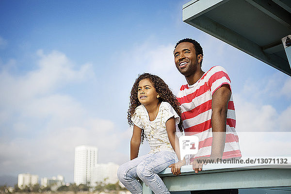 Vater und Tochter schauen weg  während sie sich am Geländer am Pier gegen den Himmel entspannen