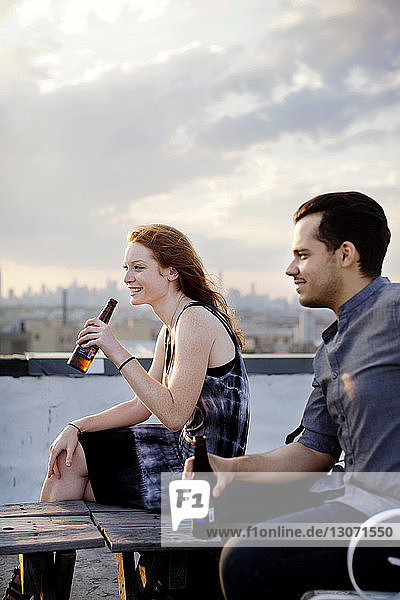 Happy couple having beer at building terrace against sky during sunset