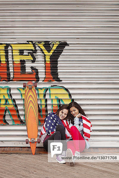 Porträt von Freunden mit amerikanischen Flaggen  die auf einem Fußweg gegen einen geschlossenen Laden sitzen