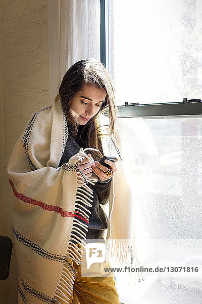 Frau benutzt Smartphone  während sie zu Hause am Fenster steht
