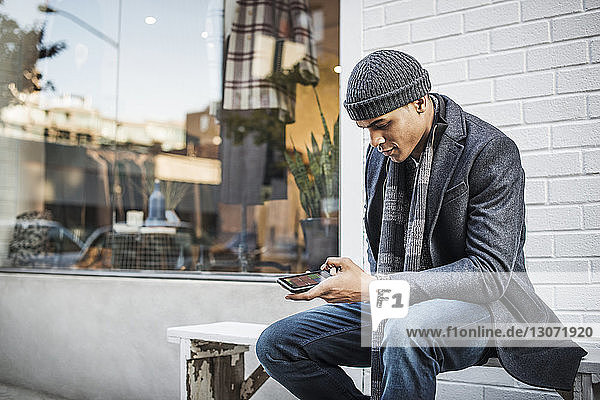 Man using mobile phone while sitting on bench against wall