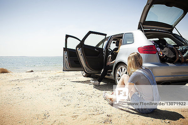 Rear view of woman wearing shoes while sitting by car at beach against clear sky