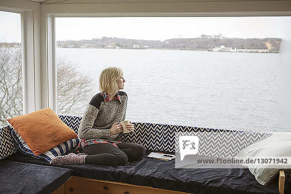 Nachdenkliche Frau trinkt im Winter Kaffee  während sie durch das Fenster auf den See schaut