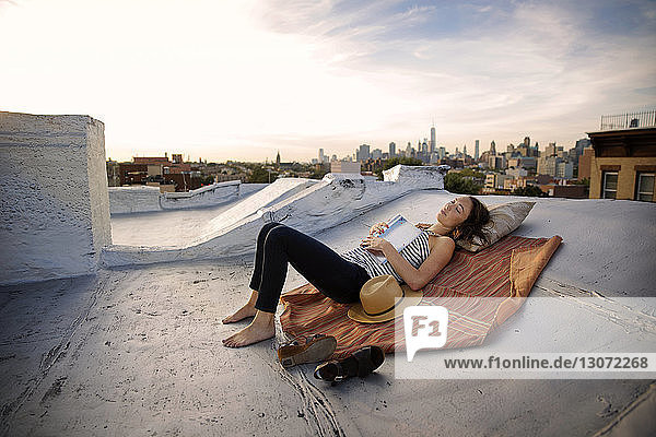 Full length of woman lying with book on building terrace against sky