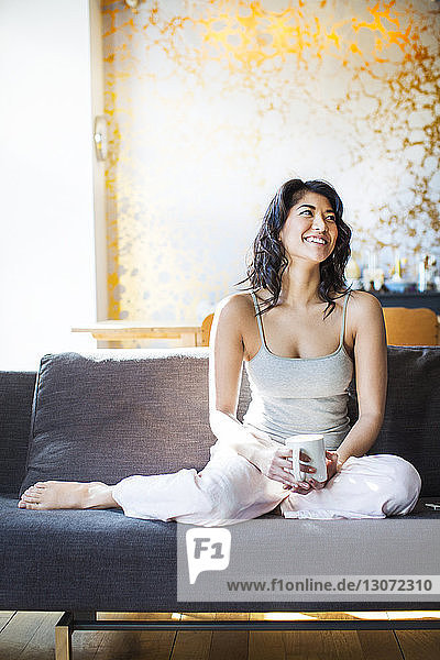 Glückliche Frau hält Kaffeetasse in der Hand und schaut weg  während sie sich auf dem Sofa entspannt