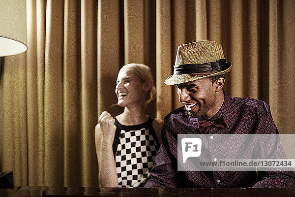 Fröhliche Frau sitzt mit einem Freund am Klavier  während sie zu Hause vor einem Vorhang sitzt