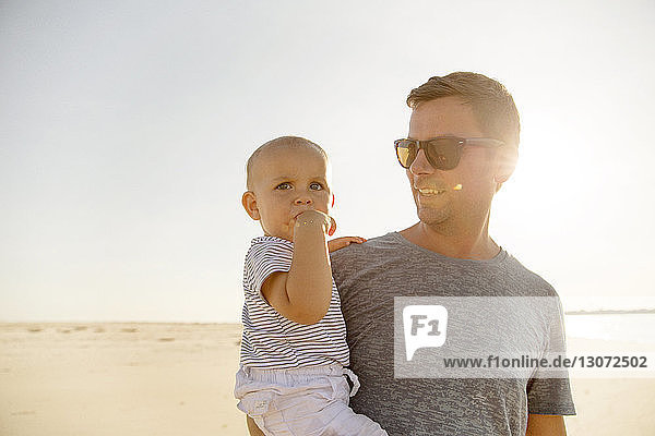 Lächelnder Vater trägt seinen Sohn  während er am Strand gegen den klaren Himmel am sonnigen Tag steht