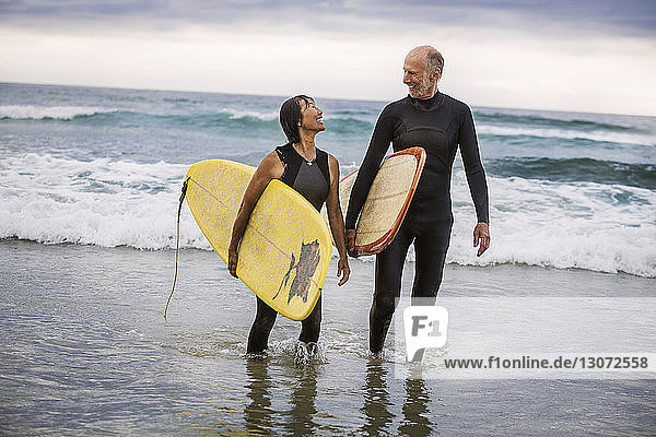 Glückliches Paar mit Surfbrettern im Gespräch beim Strandspaziergang am Ufer