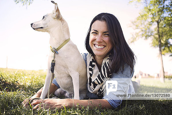 Porträt einer lächelnden Frau  die mit einem Hund auf einem Grasfeld im Park liegt