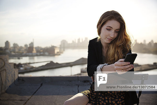 Woman using smart phone while sitting on chair against sky