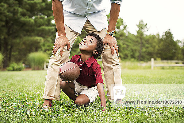 Low section of man playing football with son on grassy field in backyard