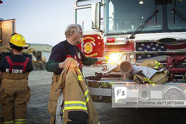 Feuerwehrmann hält Helm am Feuerwehrauto  während er seinen Kollegen im Einsatz zur Seite steht