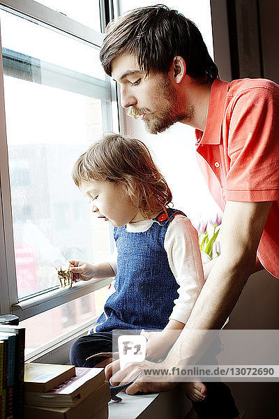 Vater sieht Mädchen an  das auf dem Fensterbrett sitzend mit Spielzeug spielt