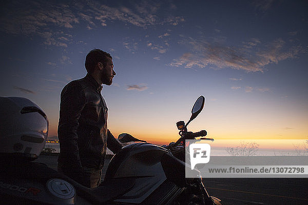 Mann steht an einem Motorrad und schaut bei Sonnenuntergang auf die Aussicht