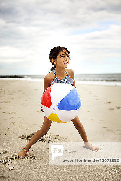 Happy girl playing with ball while standing against cloudy sky at beach