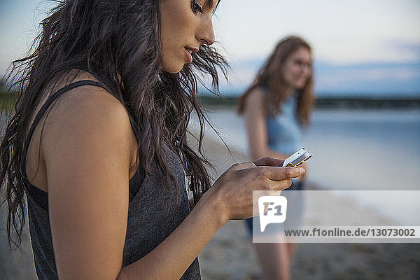 Frau benutzt Mobiltelefon  während sie mit Freundin am Strand steht