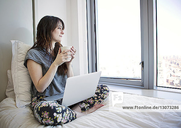Frau mit Laptop-Computer schaut weg  während sie zu Hause auf dem Bett sitzt