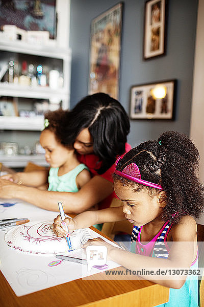 Girl drawing on paper while sitting besides mother and sister at table