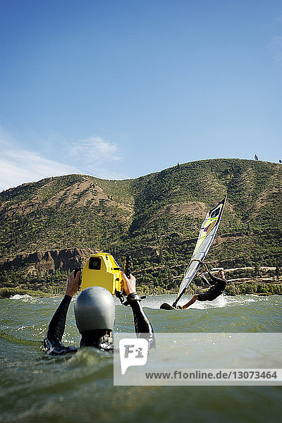 Man photographing friend windsurfing in sea against mountain