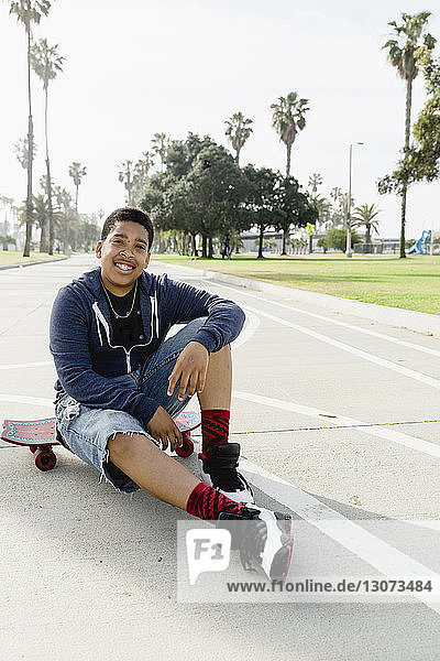 Ganzflächiges Porträt eines lächelnden Teenagers,  der im Park auf einem Skateboard sitzt