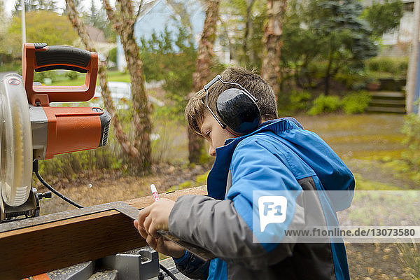 Boy wearing ear protectors while measuring wooden plank at backyard