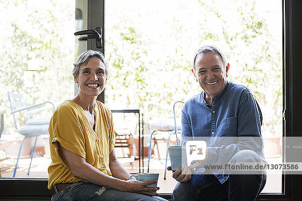 Porträt eines glücklichen älteren Ehepaares mit Kaffeebechern in der Hand  das zu Hause am Fenster sitzt