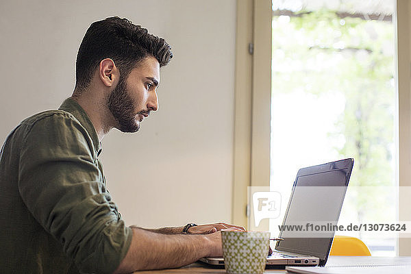 Side view of man using laptop at table by window