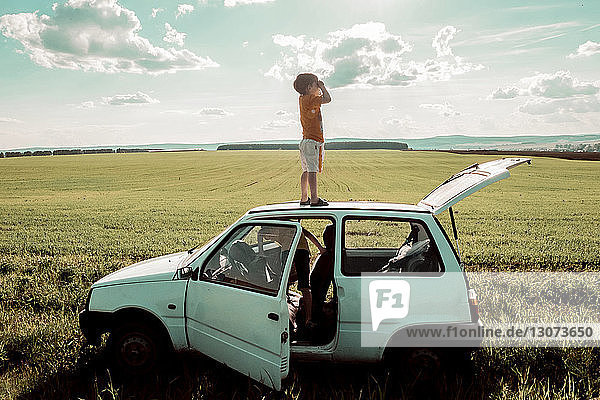 Verspielter Junge steht auf Autodach mit Bruder im Fahrzeug auf dem Feld vor bewölktem Himmel